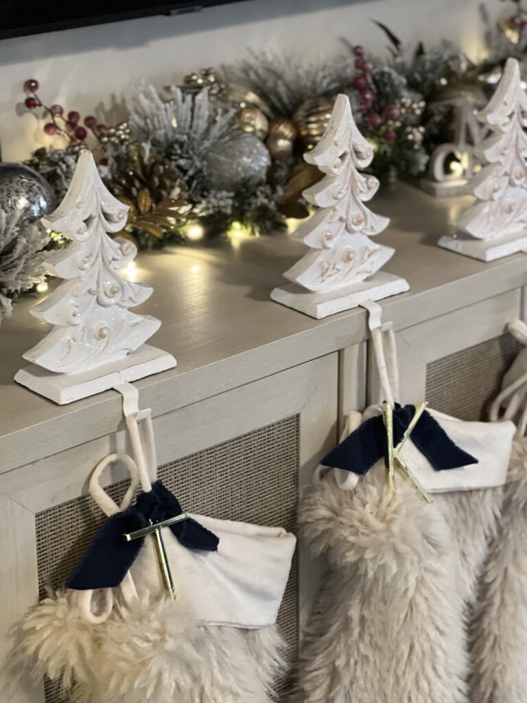 A festive mantle decorated with white Christmas tree figurines, plush stockings with navy accents