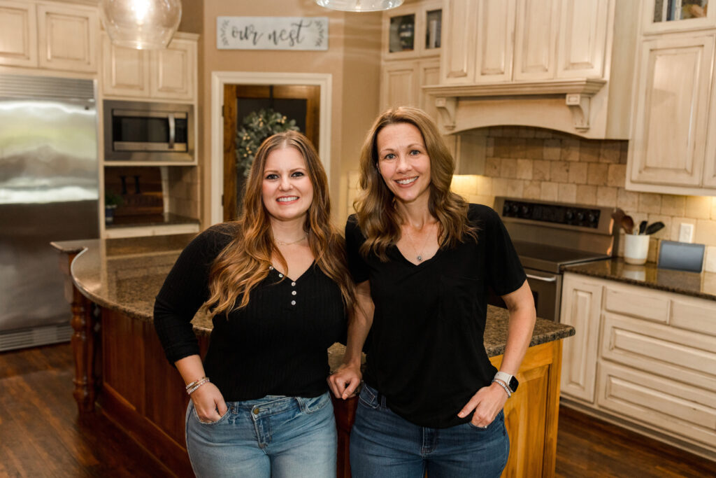 Two women smiling in a warmly lit kitchen with cream cabinets