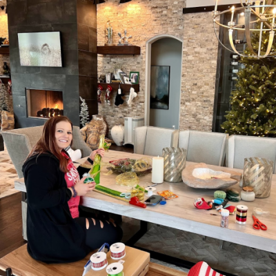 Woman crafting at a decorated table with a Christmas tree and fireplace in the background.