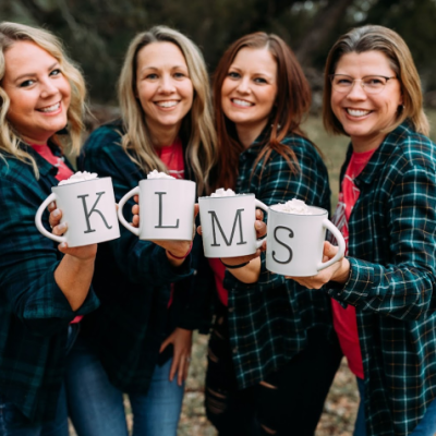 Four women smiling outdoors, holding mugs with initials